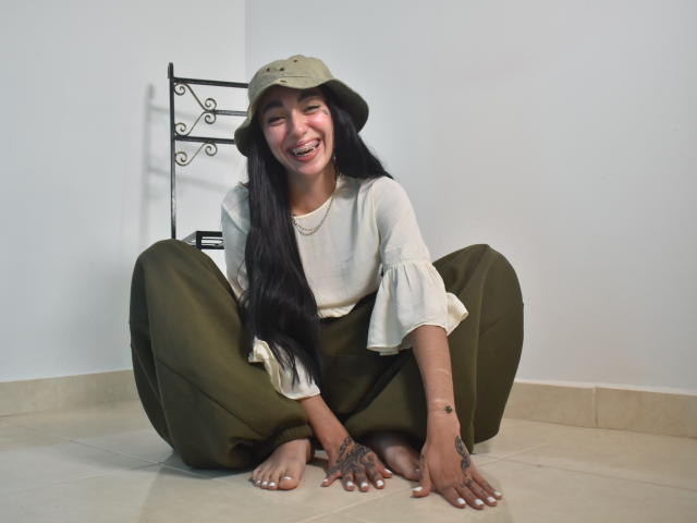 CleoZapata met lang zwart haar, lachend, met een beige hoed, een witte blouse en olijfgroene broek, zittend zonder schoenen op een tegelvloer, waarbij tatoeages op haar handen zichtbaar zijn..