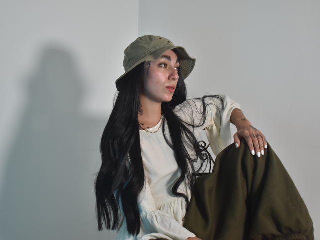 CleoZapata with long black hair, olive green bucket hat, white blouse, and green pants, sitting, looking to the side, shadow on white wall, hands on knee, subtle smile.
