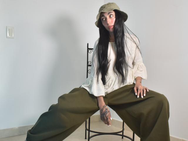 CleoZapata, avec de longs cheveux noirs, un chapeau seau vert, une blouse blanche, un pantalon olive, assise sur une chaise, une main avec des tatouages, regardant sur le côté, des ongles blancs, une peau pâle, un arrière-plan minimal..