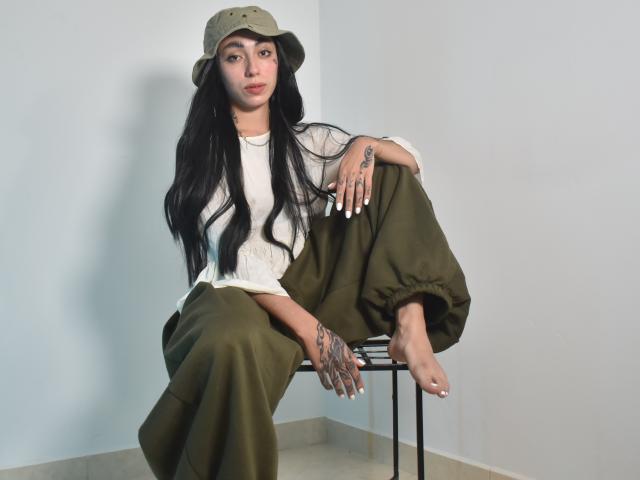 CleoZapata with long black hair, wearing a green bucket hat, white top, and olive pants, sitting on a chair, showing tattoos on hands and legs, bare feet.