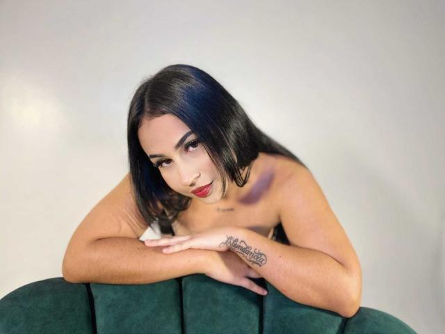 HannahBecker with black hair, red lipstick, and tattoo on her right forearm, leaning on green velvet couch, topless, looking at camera with subtle smile.