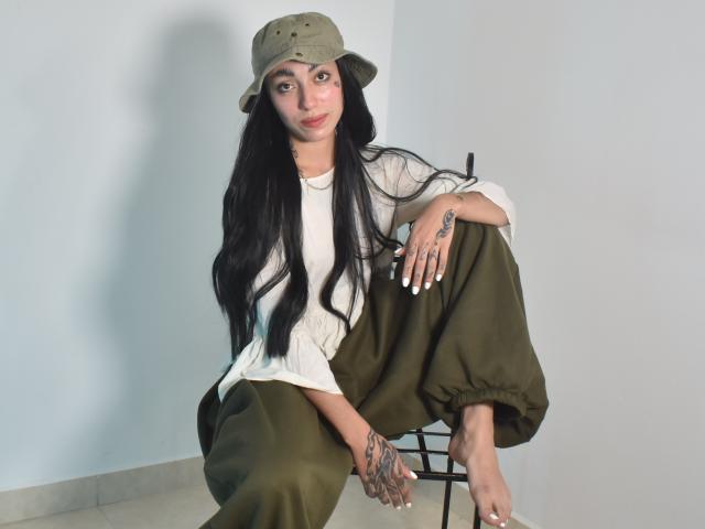 CleoZapata met lang zwart haar, olijfgroene broek, wit shirt, groene bucket hat, tatoeages op hand en voet, zittend op een stoel, witte nagellak, kijkend naar de camera..