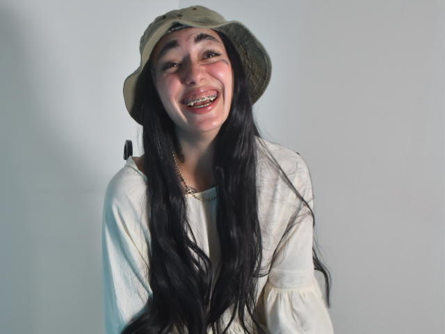 CleoZapata with long black hair, smiling, wearing a green bucket hat and white shirt, has a gold necklace, and a slightly tilted head. Her teeth are visible.