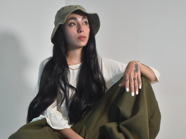 CleoZapata with long black hair, green cap, white shirt, and green pants, sitting, showing tattoo on right hand, white nails, looking up, light skin, soft light, relaxed.