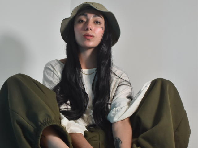 CleoZapata, con cabello negro largo, un sombrero verde oliva, una camisa blanca y pantalones verdes, está sentada con las piernas separadas, mostrando un tatuaje en su brazo derecho. Tiene una ligera expresión de ceño fruncido..