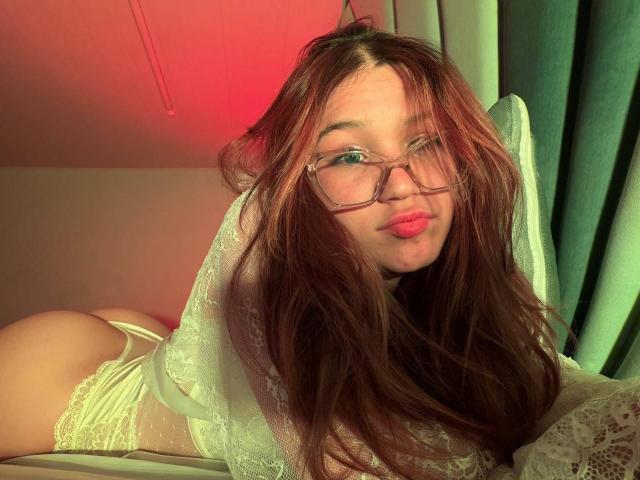 ElodieGardner with long brown hair, glasses, and pink lips, wearing white lace lingerie, lying on her stomach, showing her ass and white lace top. Red light in background.