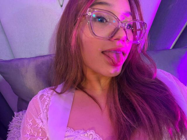 ElodieGardner with glasses, tongue out, white lace top, and pink-purple lighting, has cum on her face and chest. Her brown hair is loose, and she looks cute.
