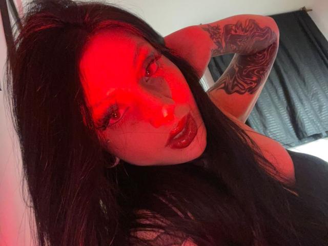 Autoportrait éclairé en rouge de Milamonty, avec des cheveux noirs, un bras tatoué, des lèvres rouges brillantes et un regard intense, portant un haut noir, mettant en valeur sa poitrine et affichant un regard sexy et séduisant..