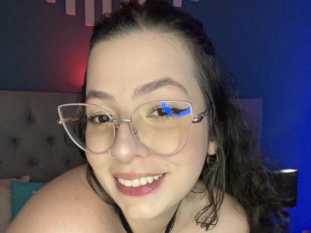 VanessaClose with glasses, smiling, nose ring, dark wavy hair, blue eyes, light skin, wearing a black strap, in a bedroom with blue and red lighting.