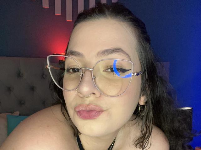 VanessaClose with glasses, nose ring, and pink lipstick, pouting her full, round lips. She has dark wavy hair, light skin, and blue eye reflection in her glasses.