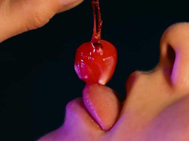 ValeriaGate's tongue is out, licking a shiny red cherry held by a hand with a sticky, glistening surface. Her nose is visible, and her skin is pinkish.