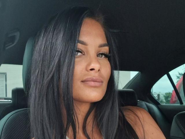 IvyMidnight, con lunghi capelli neri, sopracciglia folte e labbra piene, seduta in un'auto, che guarda di lato con un sorriso appena accennato, indossa una maglietta nera..