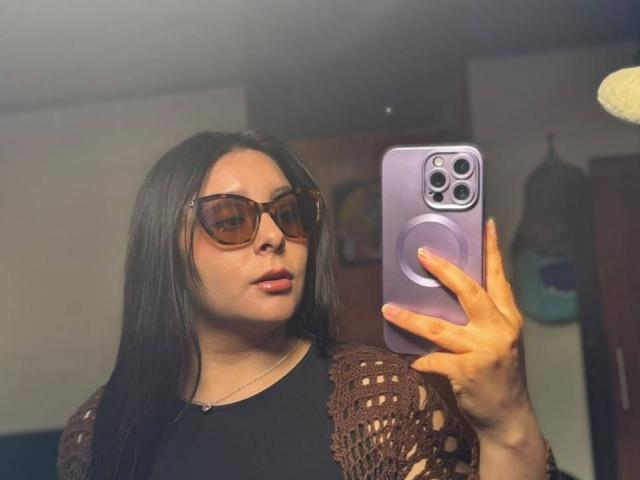 EmaJames, avec ses cheveux brun foncé, portant des lunettes de soleil et un haut en crochet marron, tenant un iPhone violet, prenant un selfie devant un miroir, montrant ses lèvres roses et sa bouche légèrement entrouverte..