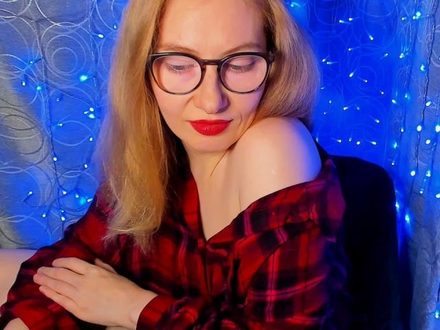 Paulina, de cabello rubio, con gafas, lápiz labial rojo y una camisa de cuadros rojos, sentada, con los brazos cruzados, mostrando un hombro descubierto, mirando hacia abajo, con luces de hadas azules en el fondo..