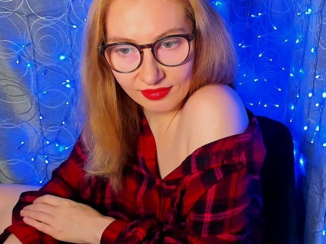 PaulinaSunny with glasses, red lipstick, and blonde hair, wearing an off-shoulder red plaid shirt, sitting with crossed arms, smiling subtly, blue fairy lights in background.