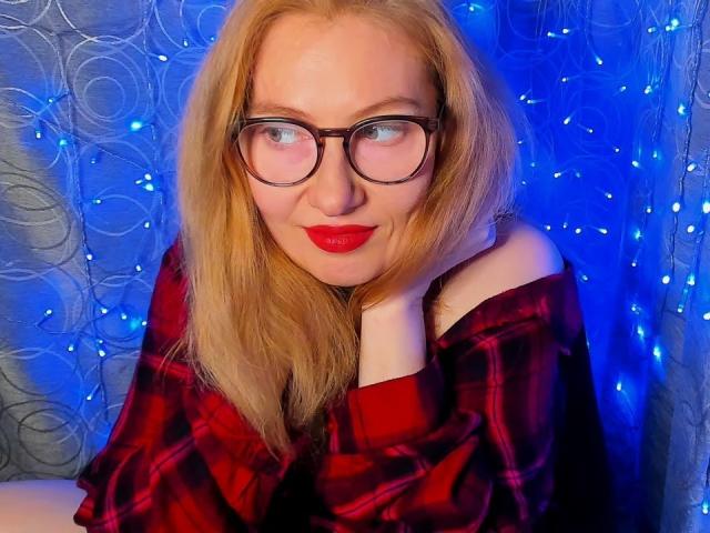 PaulinaSunny with blonde hair, glasses, red lipstick, and red plaid shirt, resting her chin on her hand, smiling softly, blue lights in the background.