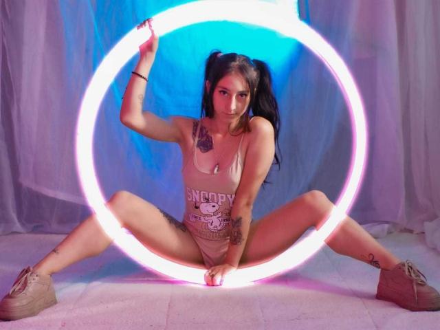 ValeriaGate with pigtails, wearing a tank top and beige sneakers, sits with legs spread, holding a neon pink ring light, showing tattoos and a cute face.