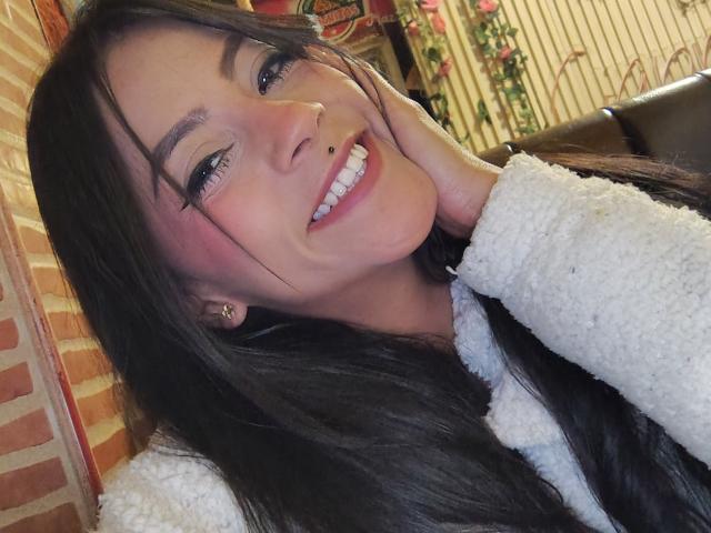 Amberv tiene el pelo largo y negro, un piercing en la nariz y está sonriendo con la mano en la mejilla. Lleva un suéter blanco y suave, y sus senos son parcialmente visibles..