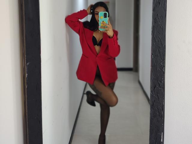 Amber se está tomando un selfie frente al espejo, vistiendo un blazer rojo, un sujetador negro y medias altas, con una pierna levantada, sosteniendo un teléfono con una funda de colores, mostrando sus pechos..