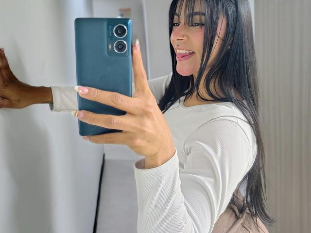 Amberv, avec ses longs cheveux noirs, sa chemise blanche à manches longues et son vernis à ongles rose, prend un selfie devant un miroir avec un téléphone bleu, montrant son visage souriant et ses fesses discrètes..