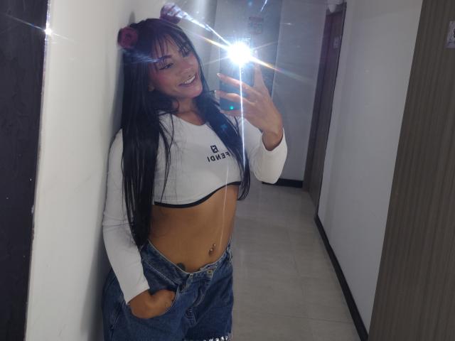 Amberv mit langen, schwarzen Haaren, weißem Crop-Top, blauen Jeans, Hände in den Taschen, lächelnd, Handy haltend, Spiegel-Selfie, leichter Lichtschein, roter Blume im Haar, Bauchnabelpiercing..