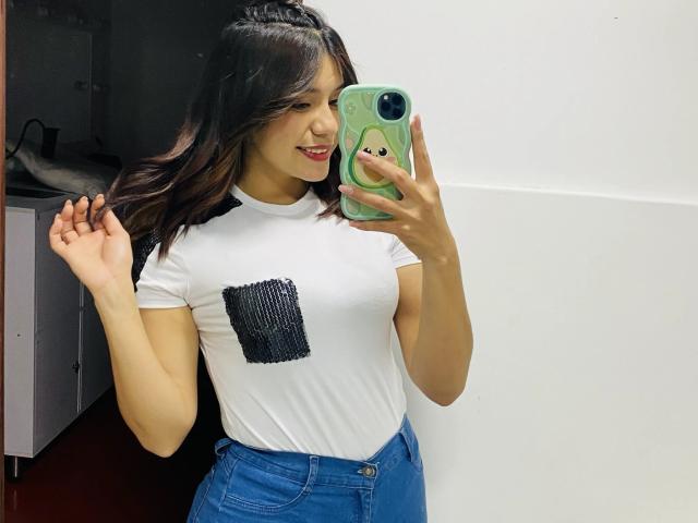 LucieMilner, com seios médios, vestindo uma camiseta branca com um quadrado preto, jeans azuis, segurando um telefone verde, sorrindo, cabelo escuro, parede branca, em pé, tirando uma selfie no espelho..