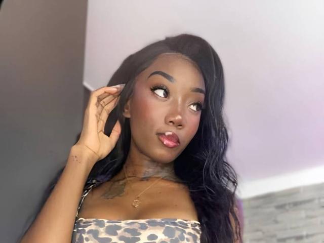 MilanCartier with dark skin, long black hair, and tattoos, wearing a leopard print top, looking to the side, hand in hair, pink lipstick, and a small necklace.