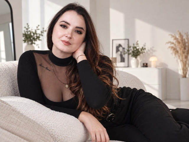 LuellaJessel with long brown hair, black sheer top showing her tits, black pants, lying on a beige couch, smiling, hand on cheek, sunlight, indoor, relaxed, sexy.
