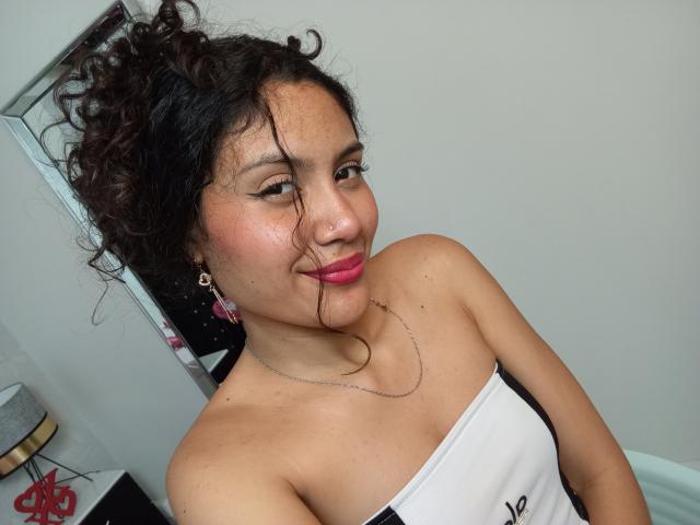 ValeryPoleth with curly dark hair, pink lipstick, and a necklace, smiling at the camera. She's wearing a strapless white top, exposing her tits. She has pink earrings.