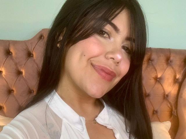 CatalinaReiyna has long black hair, brown eyes, and a cute smile with pink lipstick. She's wearing a white shirt and a small necklace, sitting on a pink tufted headboard.