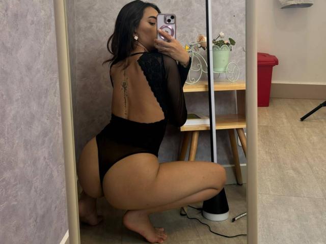 CelesteLarson is squatting, wearing a black, sheer, lace-up thong bodysuit, showing her curvy ass and tattoo on her back, taking a selfie in a mirror.