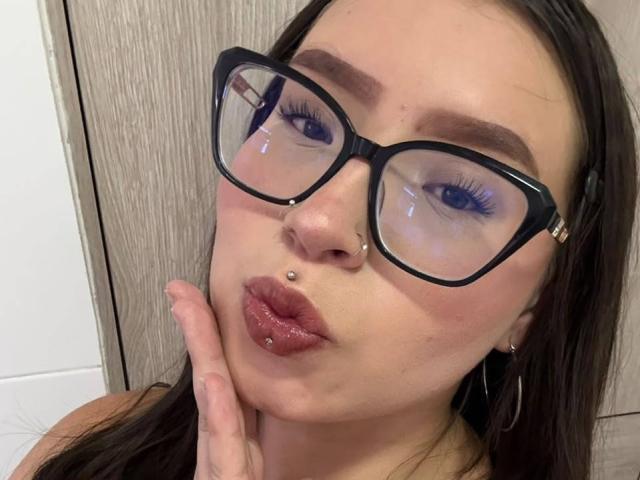 Madeleyn Rouse porte des lunettes, a un piercing au nez, un piercing à la lèvre, et une main qui touche sa joue. Elle a l'air boudeuse, avec des yeux bleus et des sourcils marrons. Ses cheveux bruns sont lâchés..