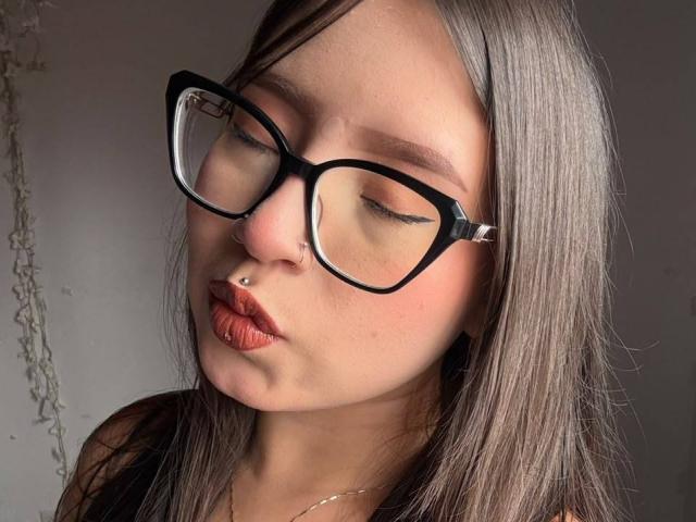 MadeleynRouse with pouty lips, brown hair, black cat-eye glasses, nose ring, and red lipstick, looking down. Boobs not visible. Soft light on face, gray background.