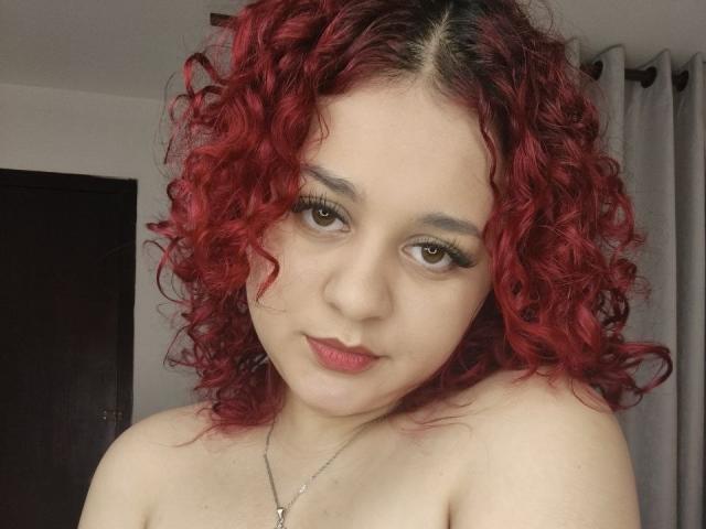 Rosalie Vittoria tiene el pelo rojo y rizado, piel clara y ojos marrones con pestañas largas. Está sin ropa en la parte superior, mostrando sus hombros desnudos y un pequeño collar de plata. Está mirando a la cámara..
