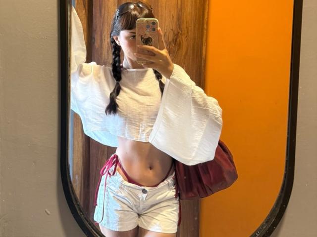 CandyyClair met gevlochten haar, een witte crop top en witte shorts met rode strikken, die een telefoon vasthoudt, een selfie in de spiegel, waarbij haar getoonde buik en kleine bil zichtbaar zijn, met een oranje muur op de achtergrond..