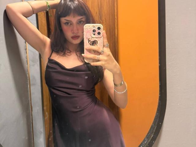 CandyyClair, con cabello castaño oscuro, vestida con un vestido de satén negro, posando frente a un espejo, sosteniendo un teléfono, mostrando sus senos, con una expresión segura, y pintalabios rojo..