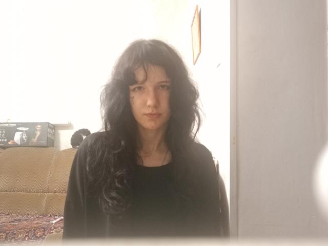 VinitaBartolomei has long, messy black hair, a serious face, and is wearing a black shirt. She stands in a sunlit room with a white wall behind her.