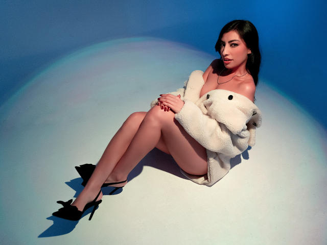 NinnaPiers with pale skin, black hair, and a cute face, sits on the floor in black heels, wearing a fluffy white jacket that's off her tits, showing her ass.