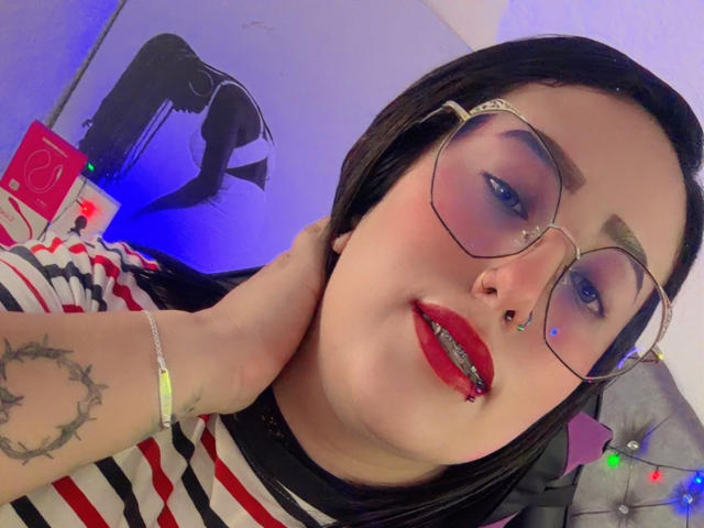 NatalieHarris with red lipstick, glasses, nose ring, and black hair, holding her neck, showing a tattoo on her arm, wearing a striped shirt. Boobs not visible.