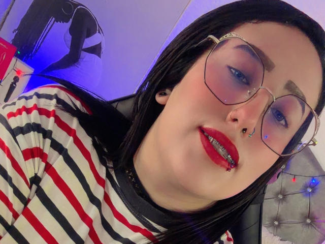 NatalieHarris with black hair, glasses, red lipstick, nose ring, and striped shirt, smiling with a slight bite on her red lip, blue eye makeup, and black ear piercings.