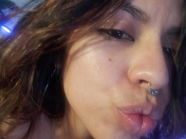 DulceBom's puffy, glossy pink lips are puckered in a kissy face, with a septum ring in her nose and slightly wavy brown hair. Her skin is light with a small mole.