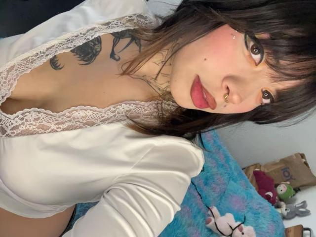 BabeSix, con cabello negro, rubor rosado y piercing en la nariz, vestida con una blusa blanca de encaje, mostrando los pezones, con un tatuaje en el cuello y brillo labial, sobre una cama con una manta azul..