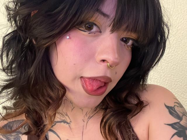 BabeSix with wavy brown hair, brown eyes, nose ring, tongue out, and tattoos on shoulders. She has a small piercing on her left cheek. Boobs not visible.