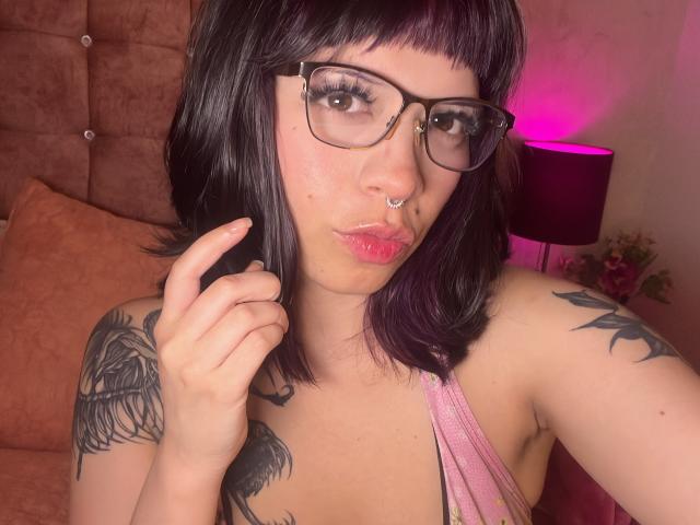 BabeSix with black hair, glasses, nose ring, pink pout, and tattoos, touching her chin, wearing a pink top, showing her tits. Pink lamp in background.