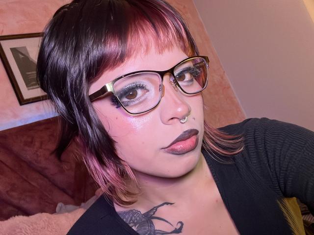 BabeSix, con capelli corti neri e rosa, occhiali, piercing al naso, rossetto nero e tatuaggio sul collo, indossa una maglietta nera e guarda la fotocamera con un leggero sorriso..