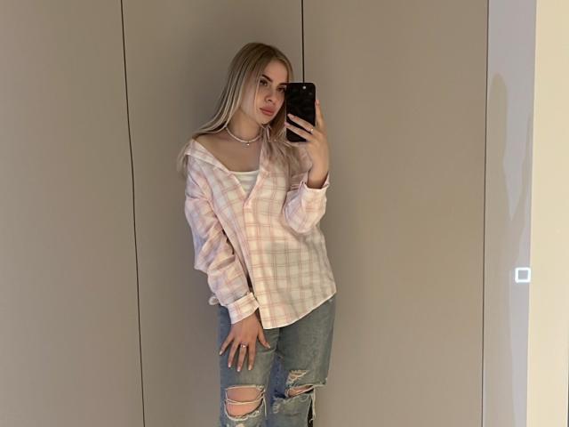 TressieBlasetti with long blonde hair, pink plaid shirt, ripped blue jeans, white choker, holding phone, looking at mirror, standing against beige wall. Boobs visible under shirt.