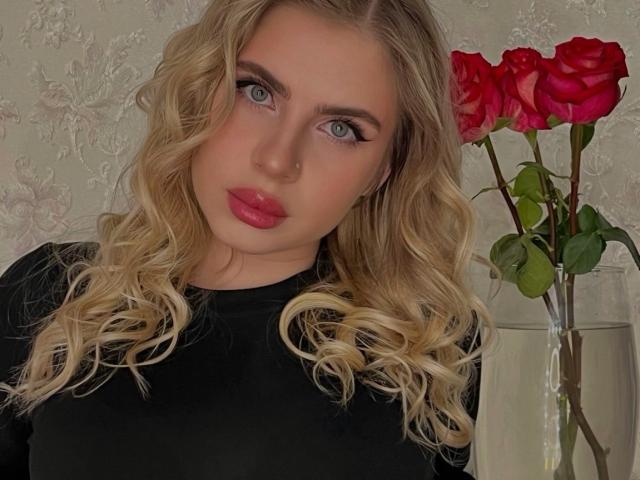 TressieBlasetti has blonde curls, green eyes, red lipstick, and wears a black top. She's holding red roses in a clear vase. Her nose is pierced.
