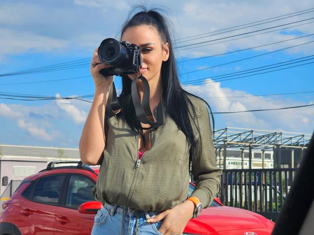CharlotteAria with long black hair, green top, and blue jeans, holding a camera to her face, standing in front of a red car, wearing a black camera strap.