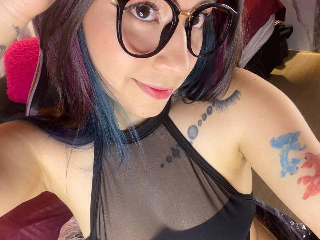 EmmaFeri, com cabelo com pontas azuis, óculos pretos e uma blusa preta transparente, mostrando tatuagens em seu braço e peito. Seus seios estão parcialmente visíveis através da blusa..