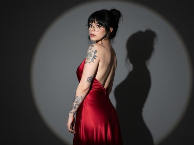 BabeSix con cabello negro recogido en un moño, gafas, vestido de satén rojo, mostrando tatuajes en los brazos y la espalda, sombra en la pared, mirando por encima del hombro, luz suave, sexy..